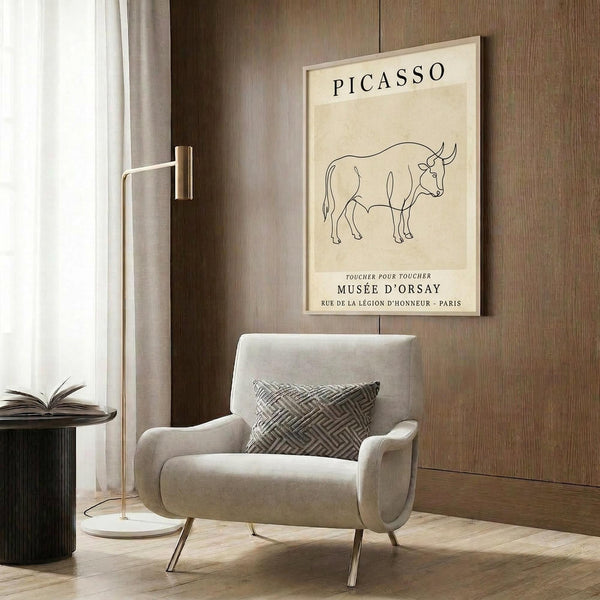 May include: Minimalist line drawing of a bull in black on cream background, framed poster on wood-paneled wall above beige mid-century modern armchair with geometric patterned pillow, gold floor lamp, and dark round side table with open book in contemporary neutral-toned room