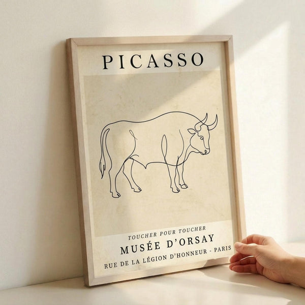May include: Framed print featuring a minimalist black line drawing of a bull on cream-colored paper, displayed against a white wall with natural lighting creating soft shadows. A hand reaches toward the wooden frame for scale reference. The artwork shows "PICASSO" at the top and "MUSÉE D'ORSAY" with French text below the bull illustration.