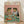 May include: Framed poster print leaning against white wall on light wood floor. Artwork shows two anthropomorphic frogs wearing sunglasses and clothing at a dinner table - one in floral Hawaiian shirt, one in striped shirt with red bow tie. They're sharing wine and food including what appears to be insects on plates. Pink background, teal table surface. Title reads "FROG PARTY" in colorful letters. Natural wood frame.