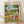 May include: Framed poster displaying two cartoon ducks doing yoga poses in a park setting. One duck balances on one leg holding a wine glass, wearing colorful floral workout clothes and sunglasses. The other duck is in downward dog pose on a pink yoga mat, wearing blue striped shirt and sunglasses with a baguette balanced on its back. Scene includes yoga blocks, wine bottle, water bottles, and snacks scattered on grass under leafy trees.