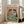 May include: Framed art print leaning against beige sofa in bright living room with large windows. The poster features two frogs wearing sunglasses and casual clothing sitting at a turquoise table with wine glasses, cheese, and insects. One frog wears a floral shirt, the other a striped shirt with red bow tie. Pink background with colorful "FROG PARTY" text at top. Light wood frame, approximately 2-3 feet tall.