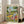 May include: Framed poster print leaning against beige armchair in bright living room with white walls and large windows. Colorful illustrated artwork shows two ducks doing yoga poses outdoors - one balancing with wine glass raised overhead, another in downward dog position. Scene includes yoga mats, wine bottle, baguettes, snacks scattered on grass under trees. Warm natural lighting creates soft shadows on hardwood floors.