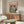 May include: Whimsical illustrated poster showing two frogs wearing sunglasses and casual clothing seated at a green checkered table with wine glasses, food, and a bottle. The artwork features a coral pink background with "FROG PARTY" text at top in colorful letters. Displayed in thin wooden frame above mid-century modern walnut credenza in minimalist living room with white curved chair, brass floor lamp, and glass-top oval coffee table.