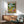 May include: Framed poster showing cartoon ducks doing yoga poses in a park setting with wine and food nearby, displayed above a mid-century modern wooden credenza with table lamp, next to a sage green upholstered armchair with wooden arms in a bright living room with white walls and natural sunlight streaming across the scene.