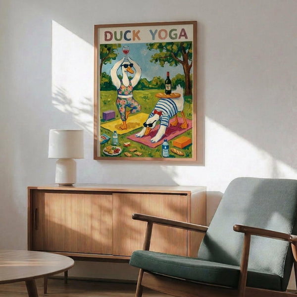May include: Framed poster showing cartoon ducks doing yoga poses in a park setting with wine and food nearby, displayed above a mid-century modern wooden credenza with table lamp, next to a sage green upholstered armchair with wooden arms in a bright living room with white walls and natural sunlight streaming across the scene.