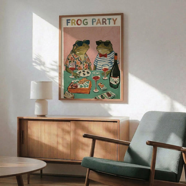 May include: Framed poster showing two frogs wearing sunglasses sitting at a green table with wine glasses, champagne bottle, and food platters against a pink background. The artwork features colorful lettering reading "FROG PARTY" at the top. Scene shows the print displayed above a wooden sideboard with white lamp, next to a mint green mid-century modern armchair with wooden arms in a bright room with natural sunlight casting shadows on white walls.