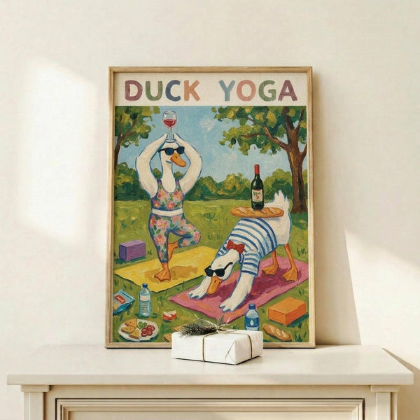May include: Framed poster showing two cartoon ducks doing yoga poses in a park setting - one duck in floral leggings holding a wine glass in tree pose, another in striped shirt doing downward dog on a pink mat. Wine bottle, baguette, cheese plate, and yoga blocks scattered around on grass. Colorful hand-painted illustration style with green trees and blue sky background. Light wood frame displayed on white wooden console table next to wrapped gift.