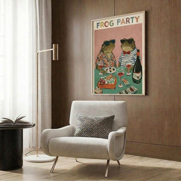 May include: Whimsical poster featuring two frogs wearing sunglasses seated at a dinner table with wine glasses, food, and a bottle on a pink and green background, displayed in light wooden frame on wood paneled wall above beige upholstered armchair with geometric pillow, alongside gold floor lamp and side table with open book.