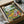 May include: Framed poster artwork in cardboard shipping box with protective wrapping. Colorful illustrated scene shows two white ducks doing yoga poses on picnic blanket in park setting. One duck holds wine glass, other balances bottle on back. Scattered food items including baguettes and cheese around blanket. Text "DUCK YOGA" at top in rainbow letters. Bright greens, blues, and warm tones throughout whimsical cartoon-style illustration.