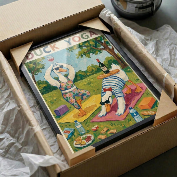 May include: Framed poster artwork in cardboard shipping box with protective wrapping. Colorful illustrated scene shows two white ducks doing yoga poses on picnic blanket in park setting. One duck holds wine glass, other balances bottle on back. Scattered food items including baguettes and cheese around blanket. Text "DUCK YOGA" at top in rainbow letters. Bright greens, blues, and warm tones throughout whimsical cartoon-style illustration.