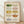 May include: Framed watercolor poster displaying ten vintage-style casserole dishes arranged in rows, featuring colorful floral patterns in green, pink, yellow, orange and blue tones against cream background, shown leaning against white wall on light wood floor with natural lighting and soft shadows
