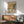 May include: Whimsical illustration poster showing two anthropomorphic dogs having tea - a poodle in floral dress and bulldog in vest - seated at table with fine china, pastries, and tiered serving stand. Artwork displayed in thin wooden frame above mid-century modern walnut credenza with white ceramic lamp, next to mint green upholstered armchair with wooden arms. Warm natural lighting creates geometric shadows on white wall.