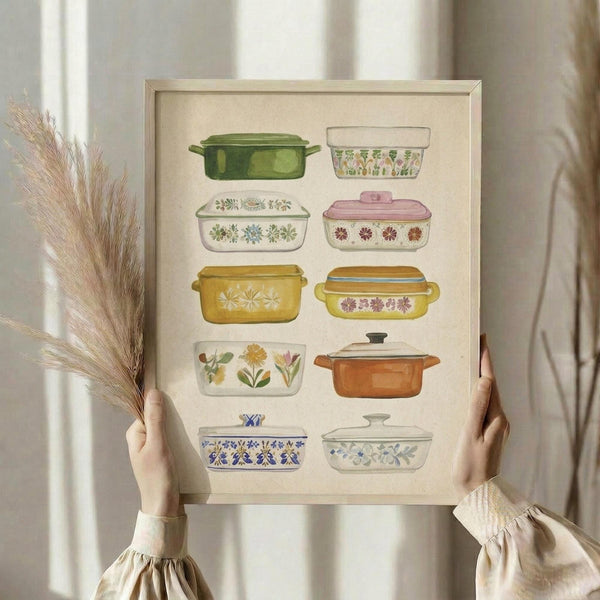 May include: Person holding framed poster print featuring watercolor illustrations of ten vintage casserole dishes and baking containers in various colors including green, yellow, orange, pink, and white with floral patterns. Light wood frame against cream background with dried pampas grass visible on left side. Person wearing beige ruffled sleeves.