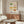 May include: Framed poster featuring watercolor illustrations of vintage casserole dishes and bakeware in various colors including green, pink, yellow, orange, and blue with floral patterns, displayed above a walnut credenza in a mid-century modern living room with white upholstered chair, glass coffee table, and copper floor lamp