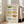 May include: Framed wall art displaying ten vintage-style casserole dishes in watercolor illustration, featuring colorful cookware with floral patterns in green, pink, yellow, orange and blue tones against cream background, shown in light wood frame leaning against white wall next to beige sofa in bright living room with large windows