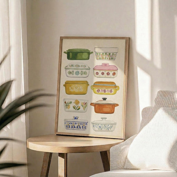 May include: Framed watercolor print showing vintage casserole dishes and baking containers in various colors including green, yellow, pink, orange, and blue with floral patterns. The artwork displays ten different retro kitchenware pieces arranged in rows against cream matting in a thin wood frame, positioned on a round wooden side table next to a beige upholstered chair with natural lighting from nearby windows.