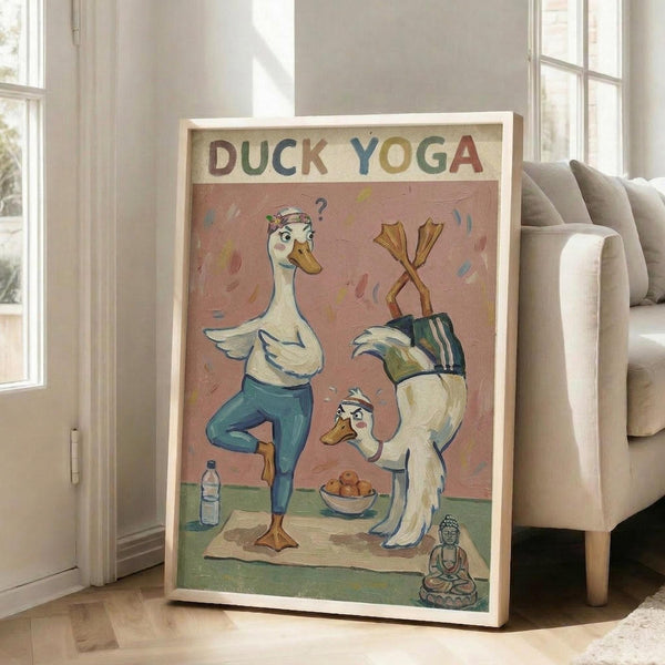May include: Framed poster with cartoon ducks doing yoga poses on pink background, displayed in bright living room corner next to beige sofa. One duck balances on one leg while another does headstand with webbed feet up. Small Buddha statue and water bottle visible on yoga mat. Colorful letters spell "DUCK YOGA" at top. Natural wood frame leans against white wall near windows.
