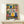 May include: A framed folk art kitchen print displayed on a light wood console table with a small wrapped gift box. The colorful illustration features stylized potted plants on shelves, hanging kitchen utensils, teapots, and mugs in vibrant blues, yellows, pinks and greens with decorative patterns. The artwork has a whimsical, hand-painted appearance with botanical elements and kitchen items arranged in horizontal bands against a cream background.
