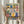 May include: Hands holding a framed folk art print depicting colorful kitchen items arranged on shelves. The artwork features decorative potted plants, hanging utensils, teapots, and cups in vibrant blues, yellows, pinks and greens with patterned borders. A bundle of dried wheat sits nearby against soft natural lighting and neutral curtains in the background.
