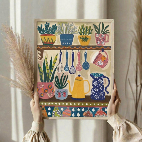 May include: Hands holding a framed folk art print depicting colorful kitchen items arranged on shelves. The artwork features decorative potted plants, hanging utensils, teapots, and cups in vibrant blues, yellows, pinks and greens with patterned borders. A bundle of dried wheat sits nearby against soft natural lighting and neutral curtains in the background.