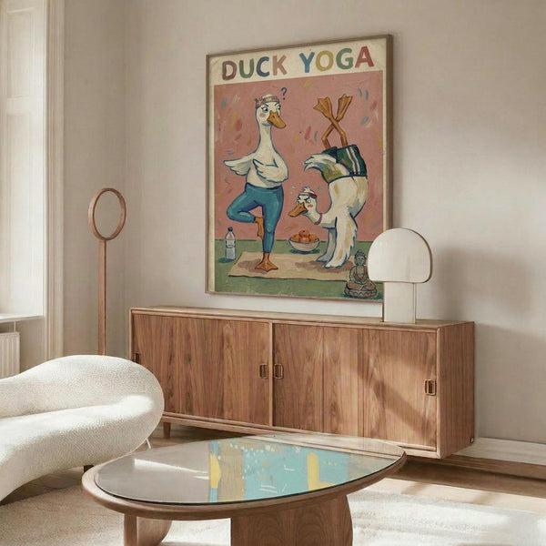 May include: Framed poster displaying cartoon ducks in yoga poses on pink background, mounted above wooden sideboard in modern living room with white curved chair, round glass coffee table, and natural lighting. The artwork shows whimsical waterfowl characters doing stretches and poses with colorful lettering at top.