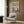 May include: Colorful folk art style poster displaying kitchen shelves with potted plants, hanging utensils, teapot, and decorative vessels in vibrant blues, yellows, pinks and greens. Mounted on wood-paneled wall above beige upholstered chair with geometric pillow. Modern brass floor lamp and dark side table with open book complete the cozy reading corner scene.
