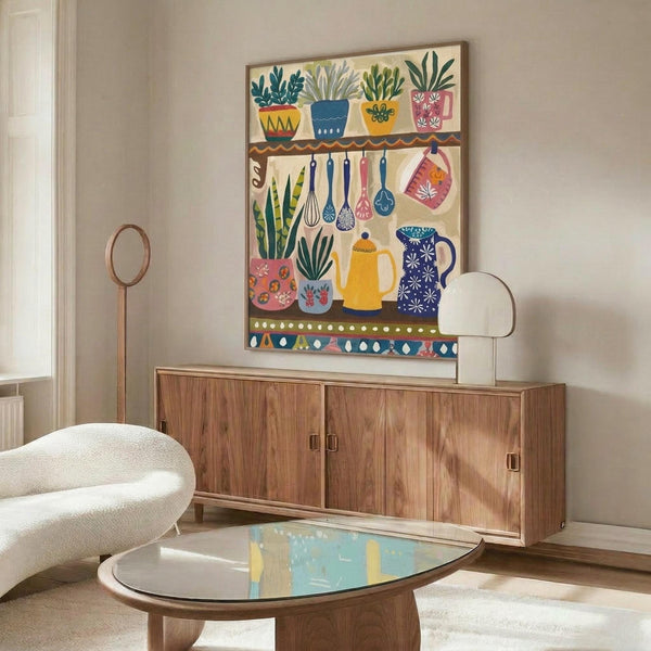 May include: Colorful folk art poster in thin wooden frame mounted above mid-century walnut credenza. Artwork features kitchen scene with potted plants on shelf, hanging utensils, ceramic vessels and teapots in bright blues, yellows, and coral tones. Modern living room setting with cream upholstered chair, glass-top oval coffee table, and white walls with natural lighting.