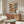 May include: Framed poster displaying cartoon cats relaxing on a tropical beach - one orange tabby lounging in a hammock wearing sunglasses and hat, another cat in Hawaiian shirt sitting nearby. Scene shows palm trees, ocean sunset, beach items scattered around. Poster hangs above mid-century modern wood credenza with white sculptural chair and glass coffee table in minimalist living room with cream walls.
