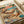 Colorful illustrated poster showing two orange tabby cats relaxing on a tropical beach. One cat wears sunglasses and a sun hat while lying in a hammock, the other wears a Hawaiian shirt and sunglasses while lounging on sand. Scene includes palm trees, turquoise ocean, pink sunset sky, scattered books, takeout containers, and a wine glass. The word "CAT NAP" appears at the top in rainbow letters. Vintage-style artwork with warm peachy and blue tones, shown flat against white paper background.