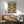 May include: Framed vintage-style poster showing orange tabby cats relaxing on tropical beach - one in hammock wearing sunglasses and hat, another in Hawaiian shirt reading. Colorful "CAT NAP" text at top. Poster hangs above mid-century modern wooden credenza with white ceramic lamp. Mint green upholstered chair with wooden arms sits nearby on wooden floor against white wall with natural sunlight streaming across scene.