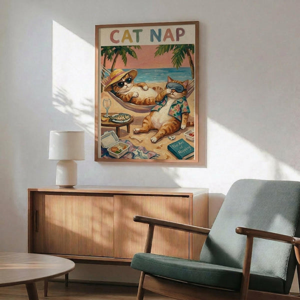 May include: Framed vintage-style poster showing orange tabby cats relaxing on tropical beach - one in hammock wearing sunglasses and hat, another in Hawaiian shirt reading. Colorful "CAT NAP" text at top. Poster hangs above mid-century modern wooden credenza with white ceramic lamp. Mint green upholstered chair with wooden arms sits nearby on wooden floor against white wall with natural sunlight streaming across scene.
