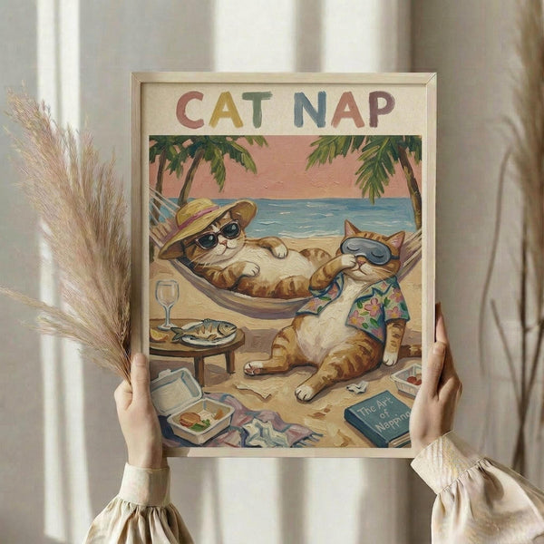 May include: Person holding framed vintage-style poster showing two orange tabby cats relaxing on tropical beach. One cat wearing sun hat and sunglasses lounges in hammock, other cat in Hawaiian shirt sits beside small table with wine glass and fish plate. Palm trees frame coral sunset sky over blue ocean. Book titled "The Art of Napping" lies on sandy beach with scattered items. Colorful "CAT NAP" text tops cream background poster in light wood frame.