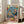 May include: Framed colorful poster leaning against white wall next to beige couch in bright living room with large windows. Artwork shows orange tabby cat wearing sunglasses and tropical shirt DJ-ing with turntables, golden retriever in black tuxedo holding cocktail shaker. Blue background with balloons, confetti, party food including cheese platter and appetizers on table below.