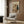 May include: Whimsical vintage-style poster showing a dachshund artist wearing sunglasses and paint-splattered smock, holding palette while painting at easel, with black cat observing nearby. Wine bottle and art supplies scattered below. Warm wood paneled wall backdrop, beige upholstered chair with geometric pillow, brass floor lamp, and round side table with open book complete the cozy reading nook setting.