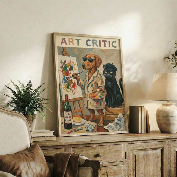 May include: Framed vintage-style poster showing a golden retriever wearing sunglasses and paint-splattered apron, holding artist palette and paintbrush, with black cat wearing beret sitting nearby. Text reads "ART CRITIC" at top. Wine bottle, easel with colorful painting, and art supplies scattered below. Displayed on rustic wooden console with books, ceramic vase, and fern plant in cozy living room setting with cream armchair.
