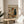 May include: Framed poster displaying a whimsical illustration of a brown dog wearing sunglasses and white paint-splattered smock, holding paintbrush and palette while standing at easel with colorful abstract painting. Black cat with beret sits nearby. Text reads "ART CRITIC" in multicolored letters. Wine bottle and art supplies scattered on floor. Poster has thin wooden frame, displayed on round wooden side table next to beige upholstered chair with white pillow. Soft natural lighting and plant