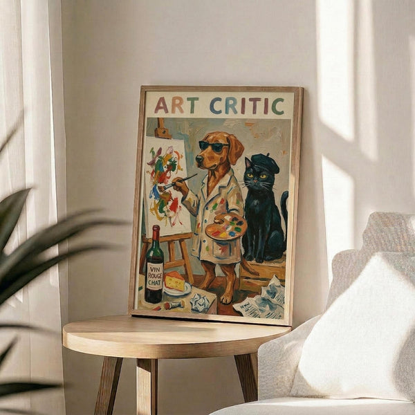 May include: Framed poster displaying a whimsical illustration of a brown dog wearing sunglasses and white paint-splattered smock, holding paintbrush and palette while standing at easel with colorful abstract painting. Black cat with beret sits nearby. Text reads "ART CRITIC" in multicolored letters. Wine bottle and art supplies scattered on floor. Poster has thin wooden frame, displayed on round wooden side table next to beige upholstered chair with white pillow. Soft natural lighting and plant