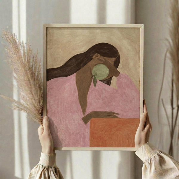 May include: Person holding framed artwork showing woman with long dark hair in pink top holding green apple, rendered in minimalist style with warm earth tones. Light wood frame against neutral background with dried wheat stems visible to the left.