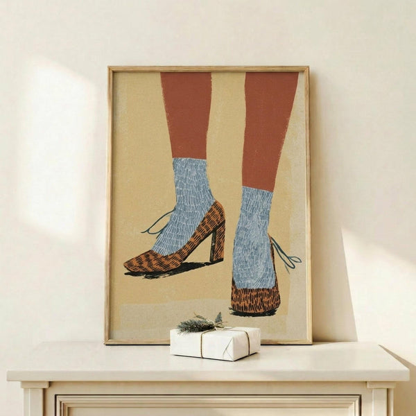 May include: Framed artwork displaying two legs wearing brown pants and blue cable-knit socks with leopard print heeled shoes, rendered in a vintage illustration style with muted colors on cream background. Natural wood frame sits on light wooden surface beside small wrapped gift with evergreen sprig against neutral wall.