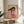 May include: Abstract portrait artwork in wooden frame showing woman with long dark hair in pink top holding green apple, displayed on round wooden side table next to beige upholstered chair with white pillow, plant leaves visible in corner, natural sunlight creating shadows on neutral wall