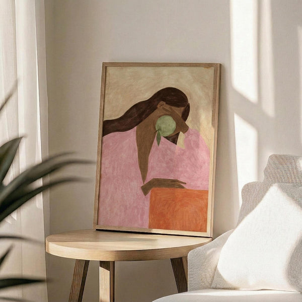 May include: Abstract portrait artwork in wooden frame showing woman with long dark hair in pink top holding green apple, displayed on round wooden side table next to beige upholstered chair with white pillow, plant leaves visible in corner, natural sunlight creating shadows on neutral wall