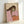 May include: Framed art print leaning against white wall showing stylized figure with long dark hair in pink top holding green apple, painted in soft muted tones with beige background, displayed in light wood frame on white surface
