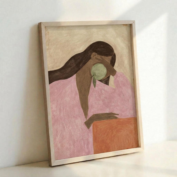 May include: Framed art print leaning against white wall showing stylized figure with long dark hair in pink top holding green apple, painted in soft muted tones with beige background, displayed in light wood frame on white surface