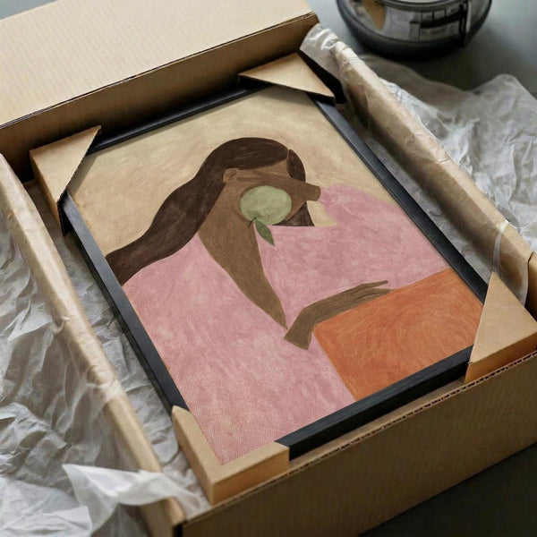 May include: Abstract art print in black frame being unpacked from cardboard box with protective tissue paper. Artwork shows stylized figure with long brown hair holding green apple, rendered in soft pink, orange and beige tones. Wooden frame corners visible alongside, suggesting assembly required. Minimalist illustration style with organic shapes and muted color palette.