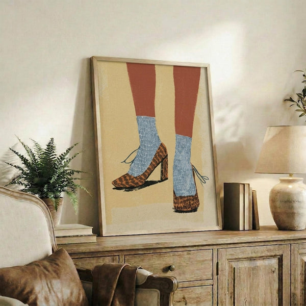 May include: Framed artwork showing a pair of legs wearing rust-colored pants, blue textured socks, and brown leopard print block heel shoes against a cream background. The vintage-style illustration sits on a wooden sideboard next to a fern plant, books, and ceramic vase in a warm, neutral-toned living room setting.