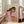 May include: Hands holding a framed abstract portrait print featuring a woman with long dark hair in pink clothing holding an orange object, painted in earthy tones with sage green accents. The artwork is displayed in a light wood frame in a bright living room with white walls, large windows, and a beige sofa with pillows.