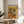 May include: Framed poster print displayed on light wood round side table next to beige upholstered chair. Artwork shows two French bulldogs in colorful Mexican attire sitting at orange table with tacos, guacamole, and tequila bottles. Yellow background with festive text. Natural lighting creates soft shadows on white wall. Plant leaves visible in corner.