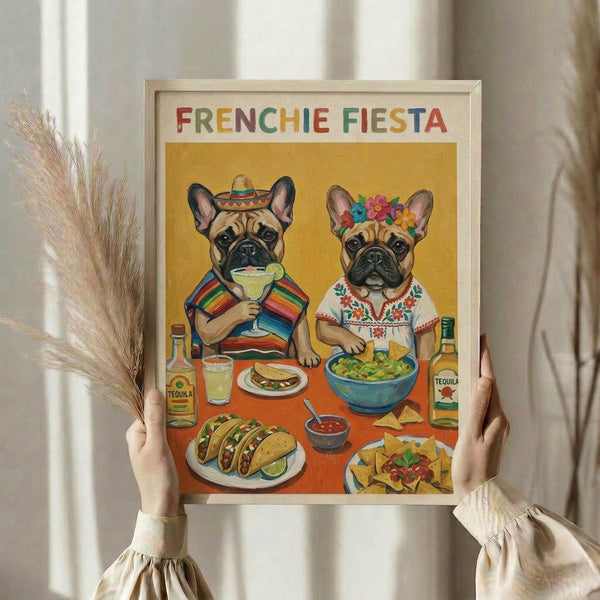 May include: Hands holding a framed poster featuring two French bulldogs in colorful Mexican attire sitting at a table with tacos, guacamole, and tequila bottles. The artwork has a warm yellow background with "FRENCHIE FIESTA" in rainbow letters at the top. Natural wooden frame, neutral beige clothing and dried pampas grass visible in soft indoor lighting.