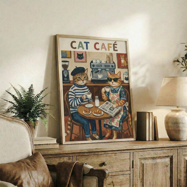 May include: Framed poster print displayed on rustic wooden sideboard showing two anthropomorphic cats sitting at cafe table with coffee cups and pastry, coffee machine in background, colorful illustrated art style with "CAT CAFÉ" text at top, surrounded by books, ceramic vase, and fern plant in cozy living room setting