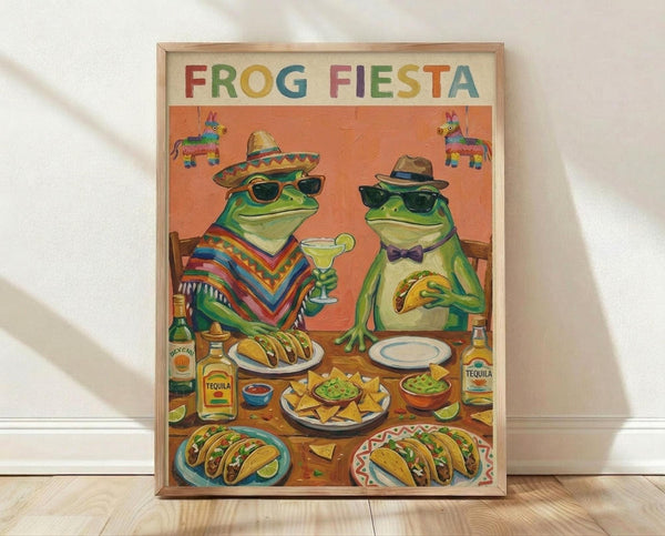 May include: Framed poster print leaning against white wall on light wood floor. Artwork shows two cartoon frogs wearing sunglasses and hats at a fiesta celebration with colorful Mexican food spread including tacos, guacamole, chips, and tequila bottles. Orange background with hanging piñatas. One frog wears a striped poncho and sombrero holding a margarita, the other wears a fedora and bow tie holding a taco. Vibrant colors throughout with wooden frame.